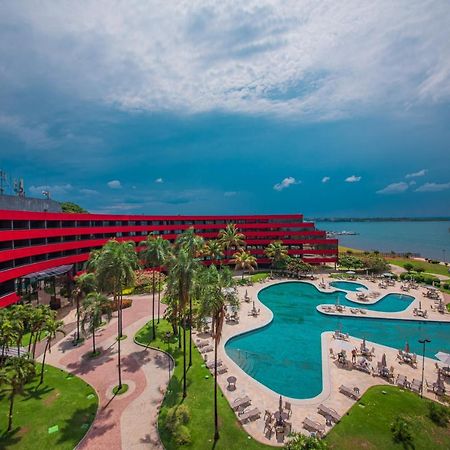 Royal Tulip Brasilia Alvorada Hotell Exteriör bild The photo shows a modern hotel building with a striking red exterior, surrounded by lush greenery. In the foreground, there is a large, creatively shaped swimming pool, with lounge chairs arranged around it. The backdrop features a body of water, pos
