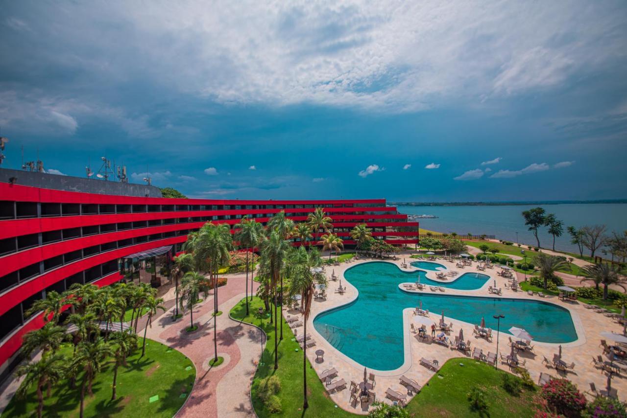 Royal Tulip Brasilia Alvorada Hotell Exteriör bild The photo shows a modern hotel building with a striking red exterior, surrounded by lush greenery. In the foreground, there is a large, creatively shaped swimming pool, with lounge chairs arranged around it. The backdrop features a body of water, pos