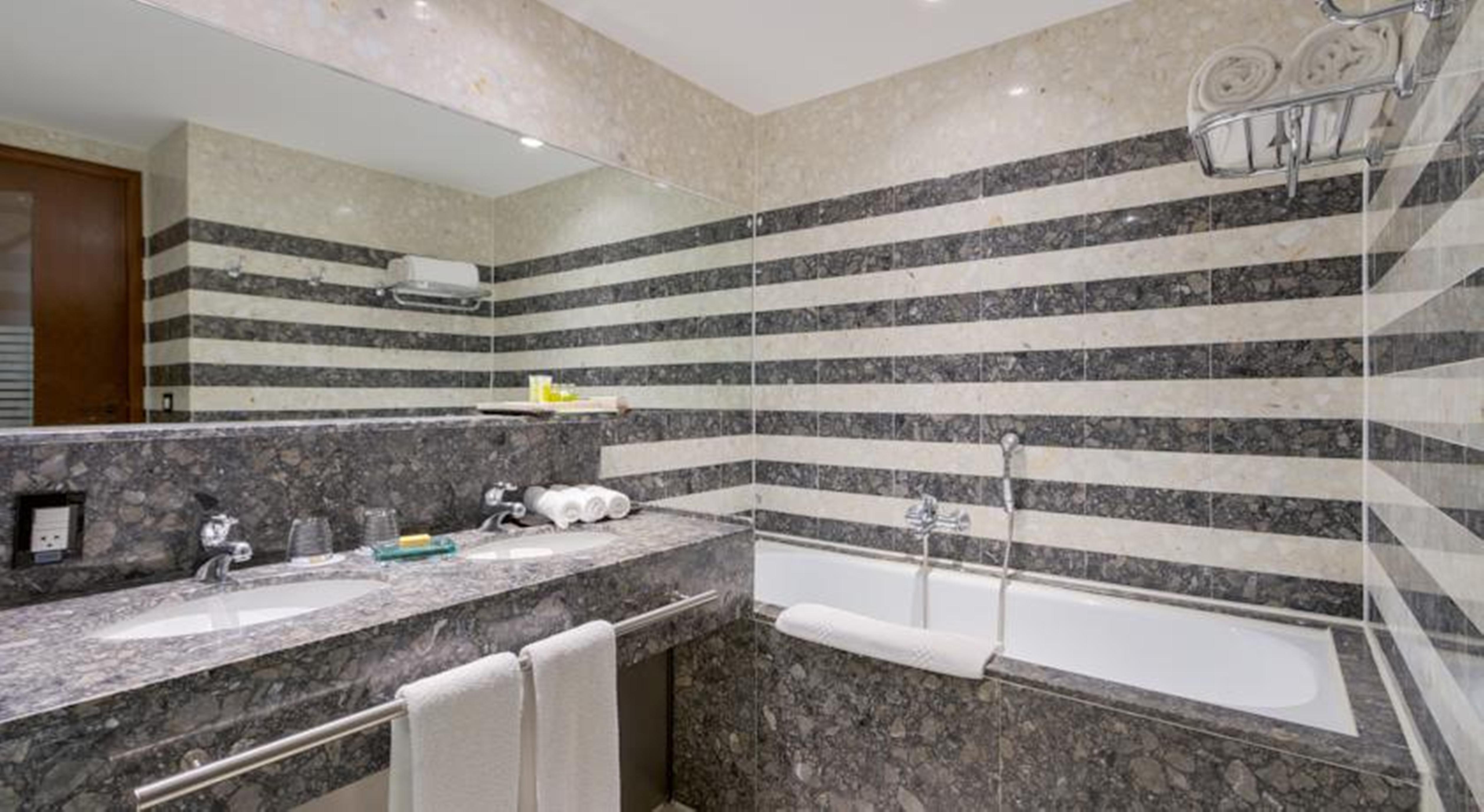 Royal Tulip Brasilia Alvorada Hotell Exteriör bild The photo shows a modern bathroom featuring a combination of marble and stone finishes. The walls are decorated with horizontal stripes in various shades of gray and cream. There is a bathtub with a white exterior, and a showerhead is mounted on the 