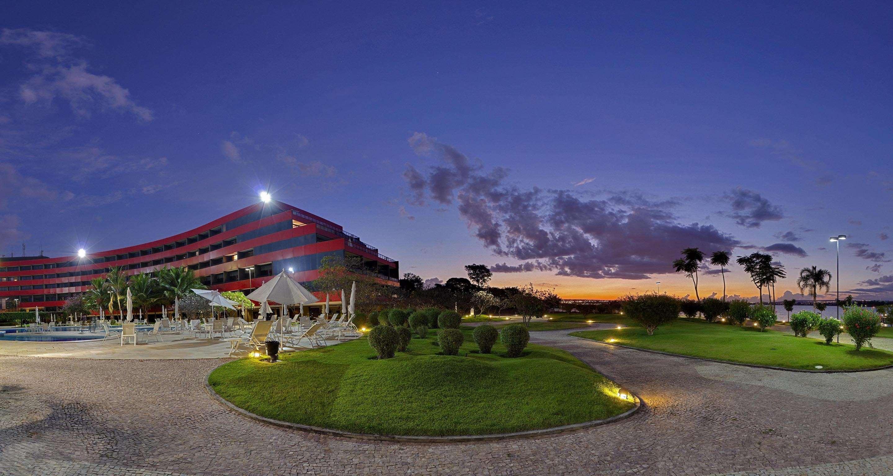 Royal Tulip Brasilia Alvorada Hotell Exteriör bild The photo depicts a modern building, possibly a hotel or resort, with a striking red exterior. It is set in a landscaped area that features well-maintained greenery and ornamental plants. In the foreground, there are lounge areas with umbrellas, sugg