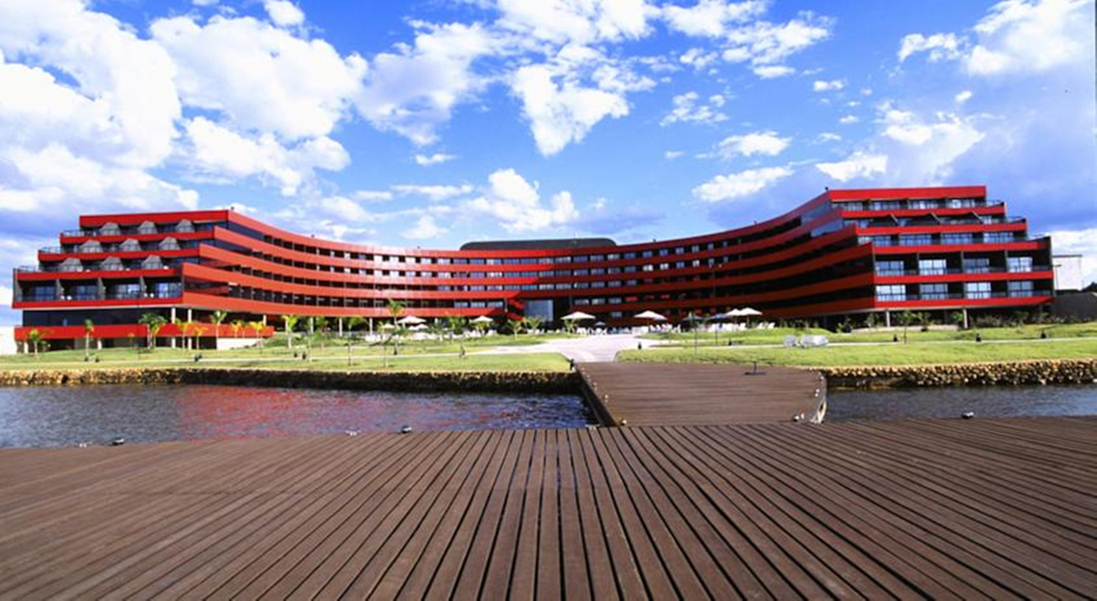 Royal Tulip Brasilia Alvorada Hotell Exteriör bild The image shows a modern, curved building with a striking red facade, situated near a body of water. The architecture features multiple levels with large windows that create a sleek and contemporary appearance. In front of the building, there is a wo