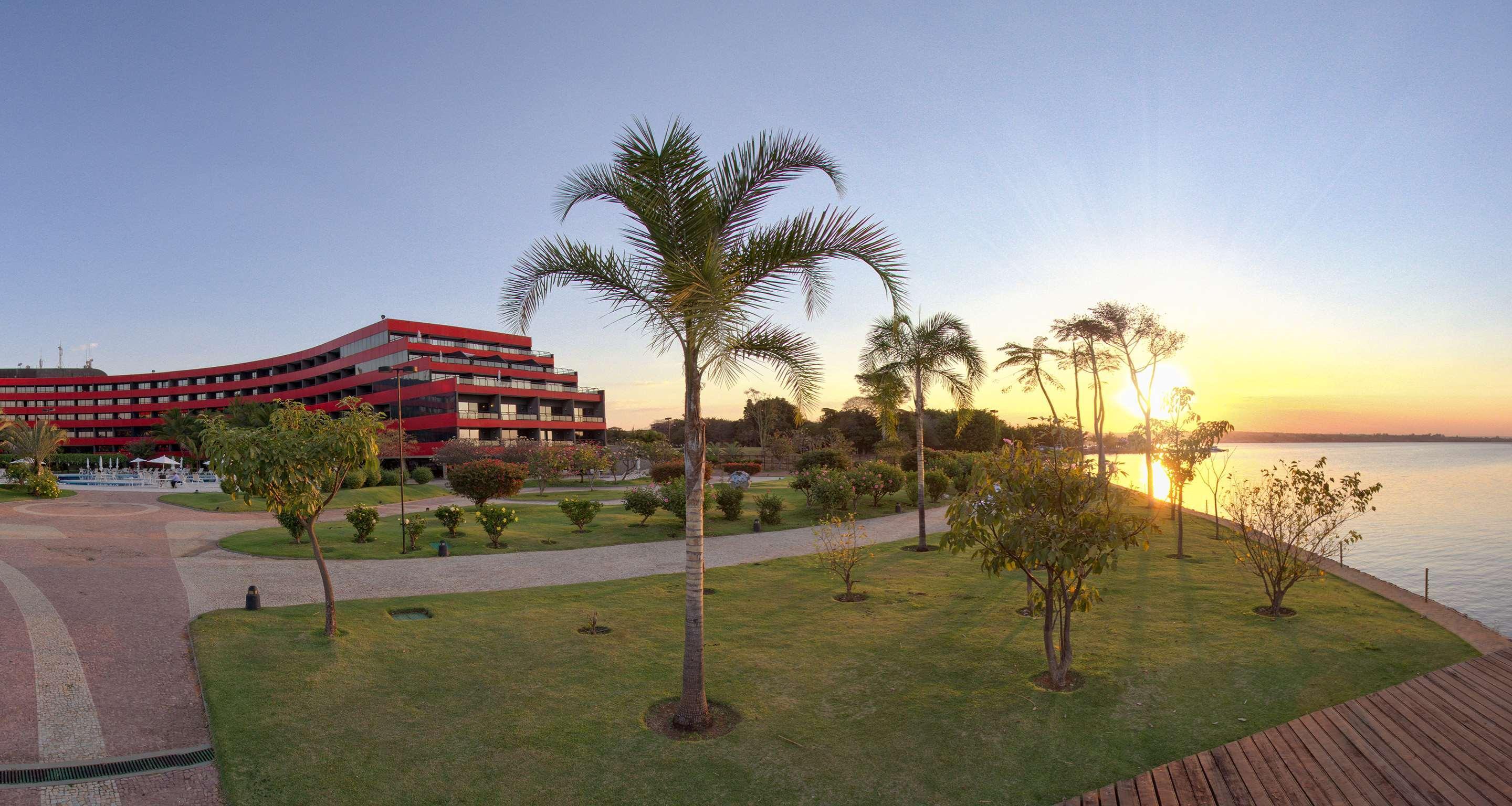 Royal Tulip Brasilia Alvorada Hotell Exteriör bild The photo depicts a scenic view featuring a red multi-story building alongside a serene waterfront. In the foreground, there are several palm trees and lush green lawns, creating a tropical atmosphere. The sun is setting in the background, casting wa