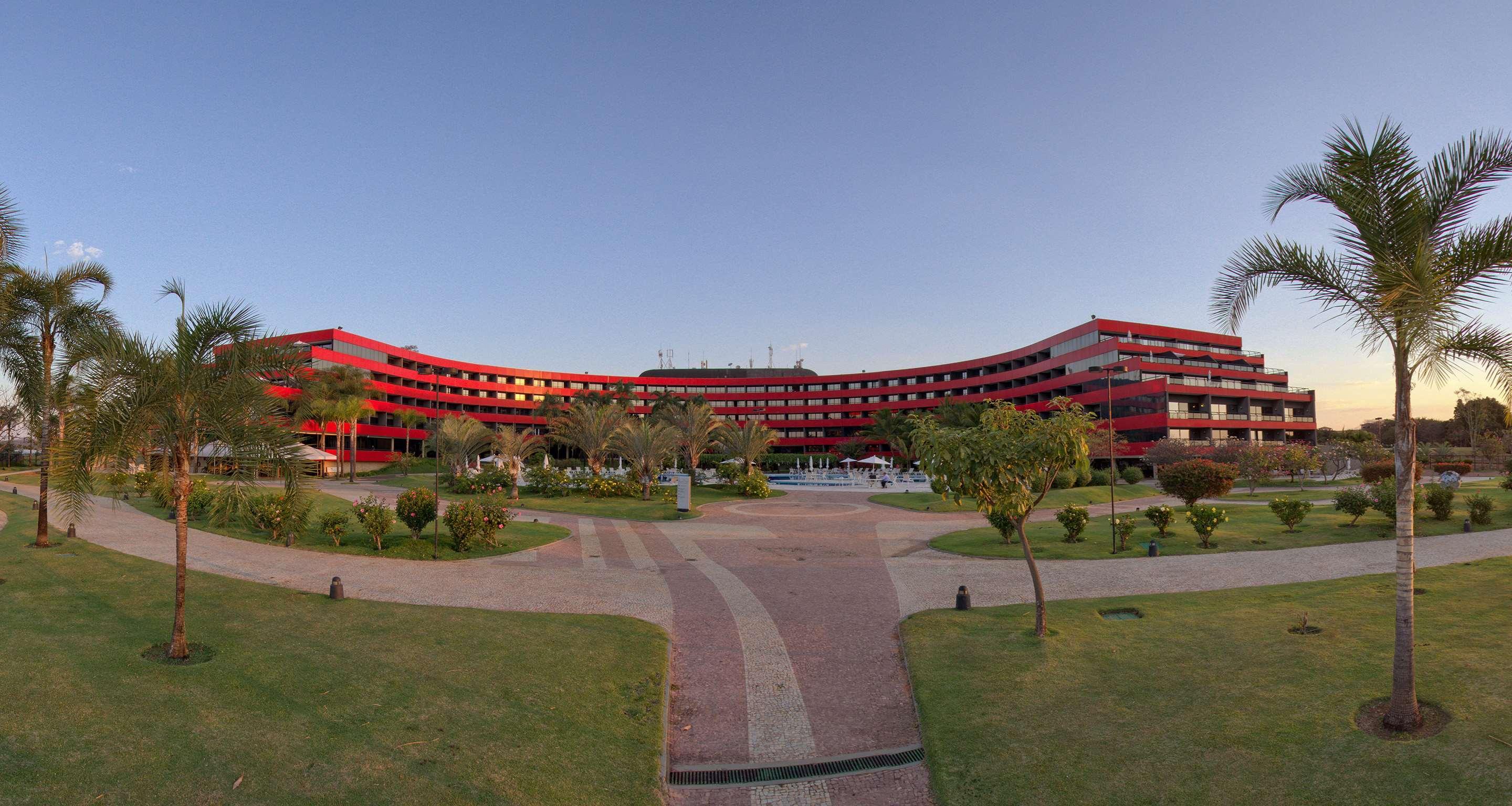 Royal Tulip Brasilia Alvorada Hotell Exteriör bild The photo shows a large, curved building with a vibrant red exterior. It appears to be a hotel or resort, surrounded by well-maintained gardens and green spaces. The landscape includes manicured lawns, small trees, and pathways, suggesting a pleasant