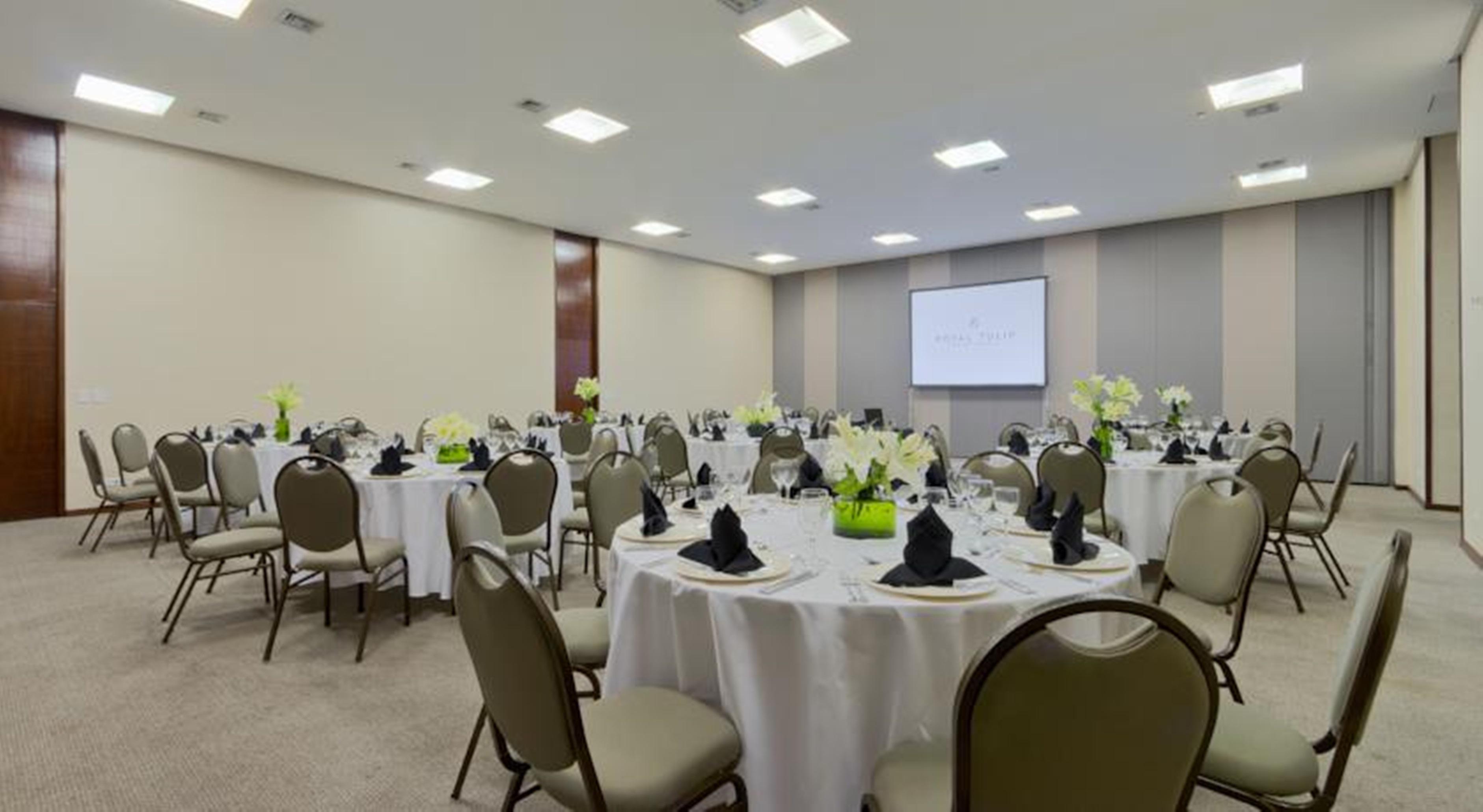 Royal Tulip Brasilia Alvorada Hotell Exteriör bild The photo shows a banquet or conference room set up with multiple round tables covered in white tablecloths. Each table is arranged with black napkins and centerpieces featuring flowers, likely in shades of green and white. The room has beige walls a