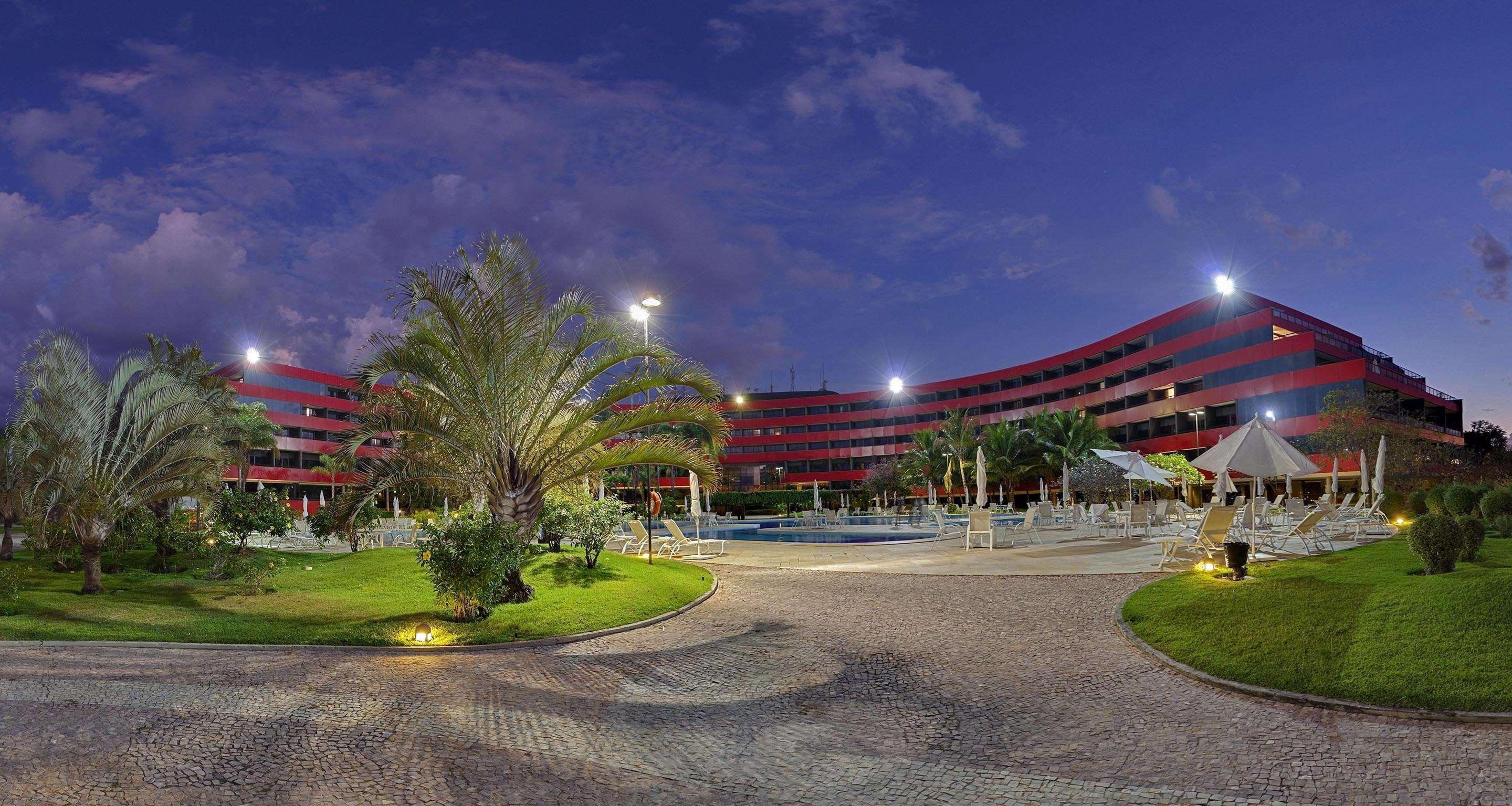 Royal Tulip Brasilia Alvorada Hotell Exteriör bild The photo shows a panoramic view of a hotel or resort complex, characterized by its modern architectural design with smooth, curved lines and a vibrant red color. It’s set in a landscaped area featuring palm trees and green grass. The surroundings in