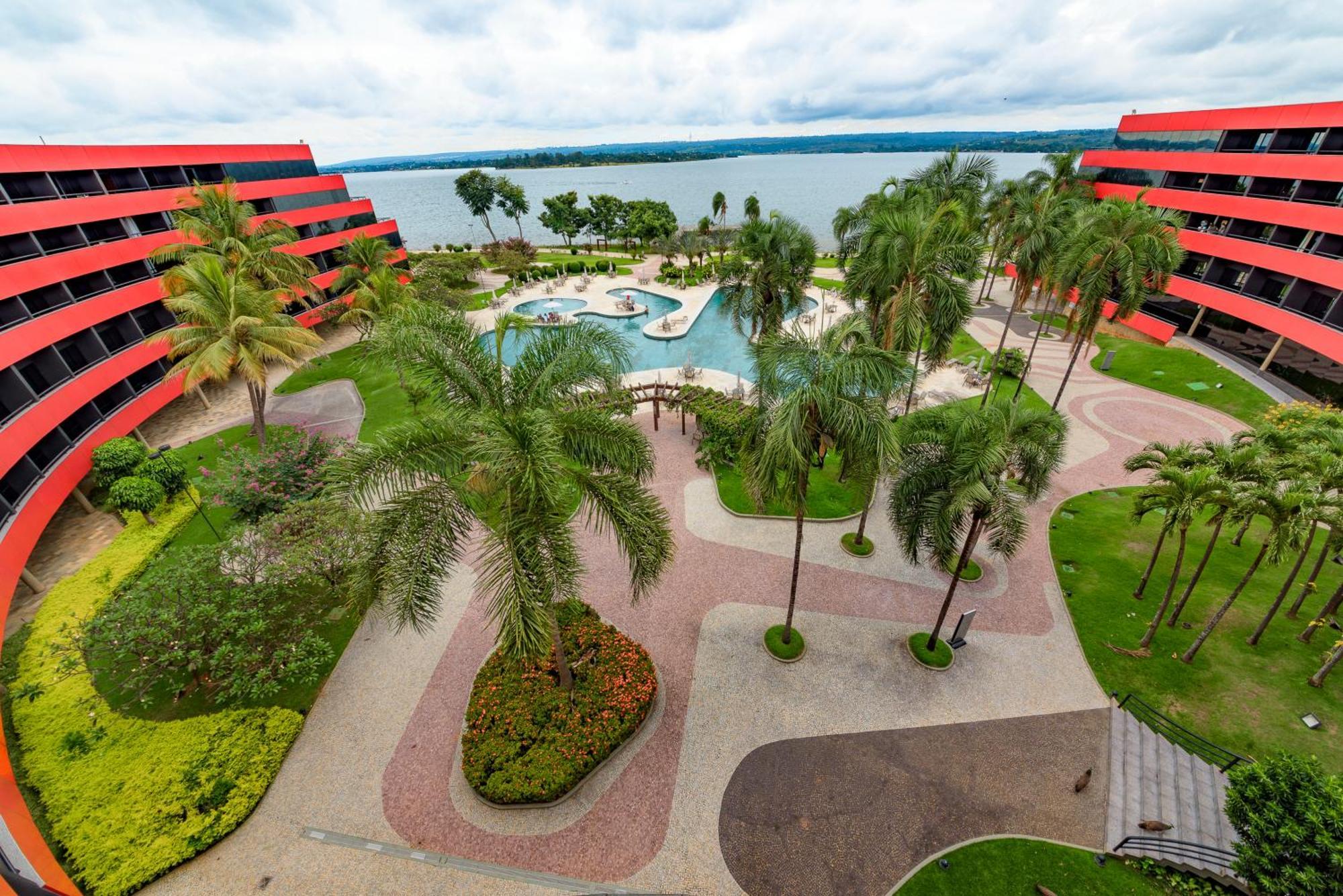 Royal Tulip Brasilia Alvorada Hotell Exteriör bild The photo shows a scenic view of a courtyard area in a residential complex. There are multiple red buildings surrounding a pool area, which is surrounded by palm trees and landscaped gardens. The pool features a curvy design, and there are pathways m