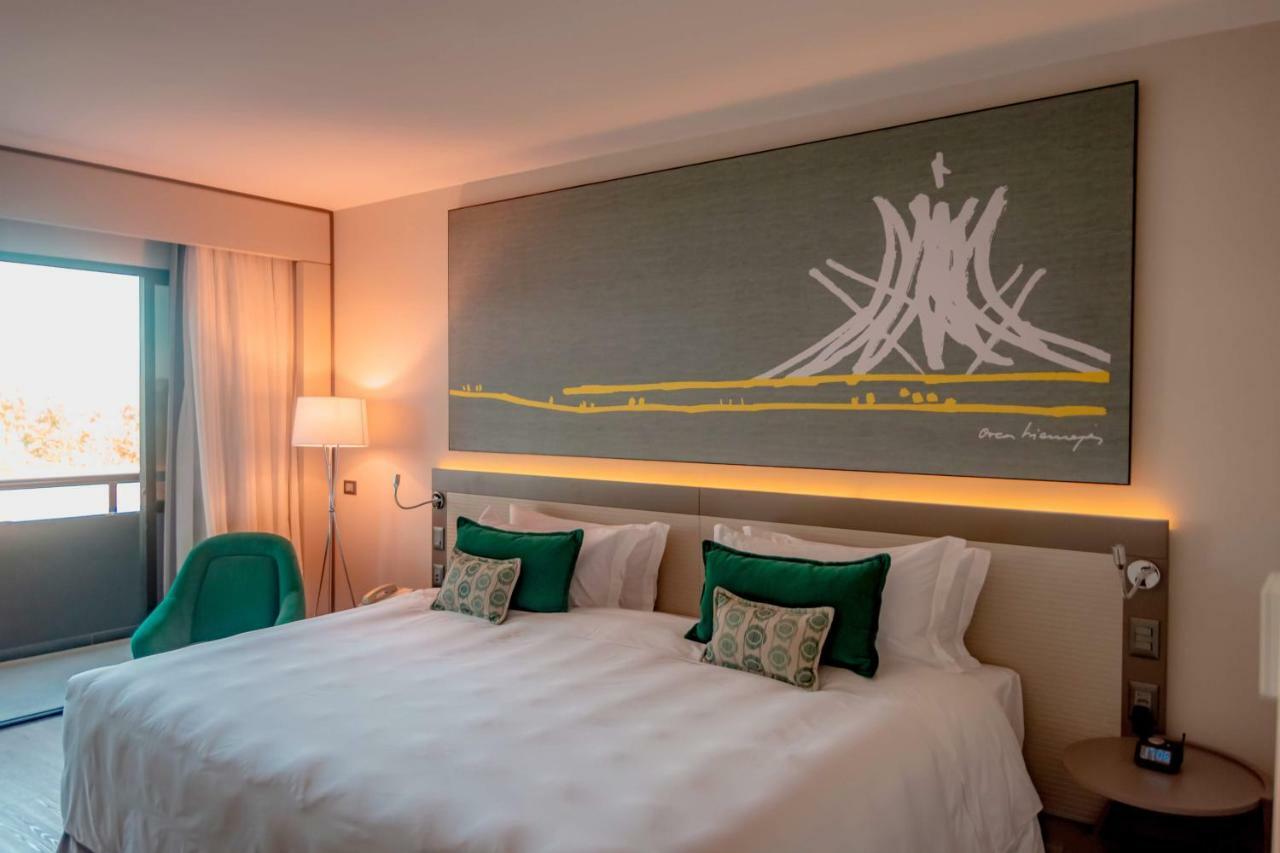 Royal Tulip Brasilia Alvorada Hotell Exteriör bild The photo shows a modern hotel room featuring a large bed with white bedding and two green accent pillows. Above the bed, there is a large piece of artwork depicting a stylized mountain or volcanic shape with yellow accents. The room has a contempora